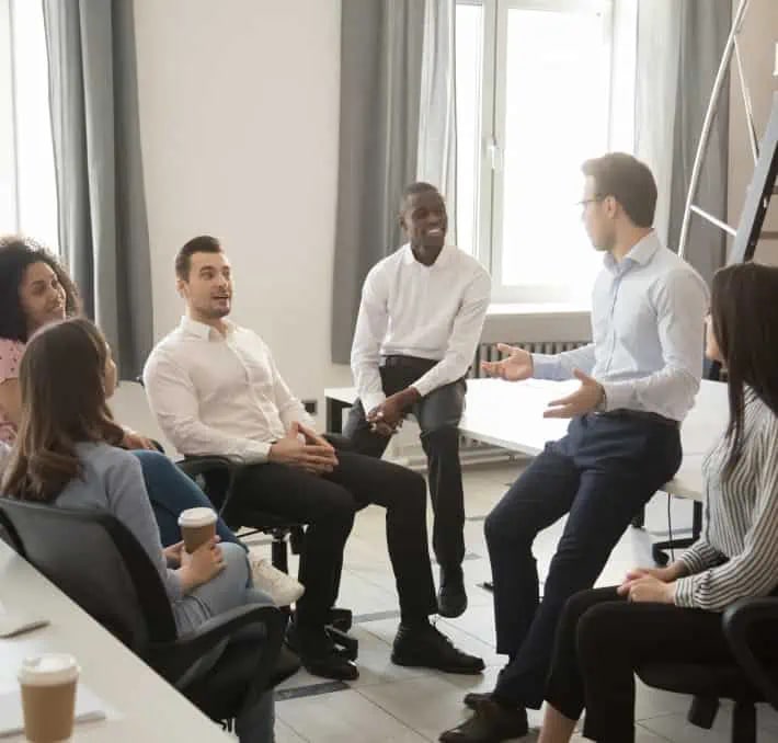 A group of six people sit in an office, engaged in a discussion. One man gestures while speaking, and others listen attentively. Coffee cups are visible on the table and in hands.