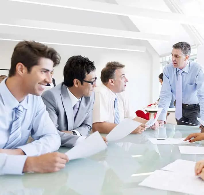 Five people in business attire are seated around a glass conference table, holding papers and having a discussion in a bright office setting.